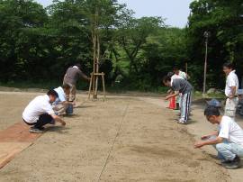 地面にしゃがみ込み、ジャージ姿の大人たちが芝生化する区画へ種蒔きをしている写真