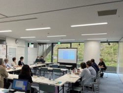 3名がモニターの横で説明をし、コの字型に並べられた長机に座っている8名がモニターに表示されている報告内容に注目している様子を撮影した写真