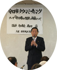守口市タウンミーティングとテーマ書かれたが紙が壁に掲示された前で1人のスーツ姿の男性がマイクを持ち立ちながら話をしている写真