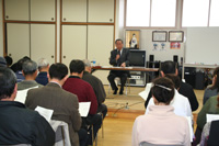 前方に1人の男性が座りながら説明をしていて、向かい合わせに参加者の人達が座っている写真