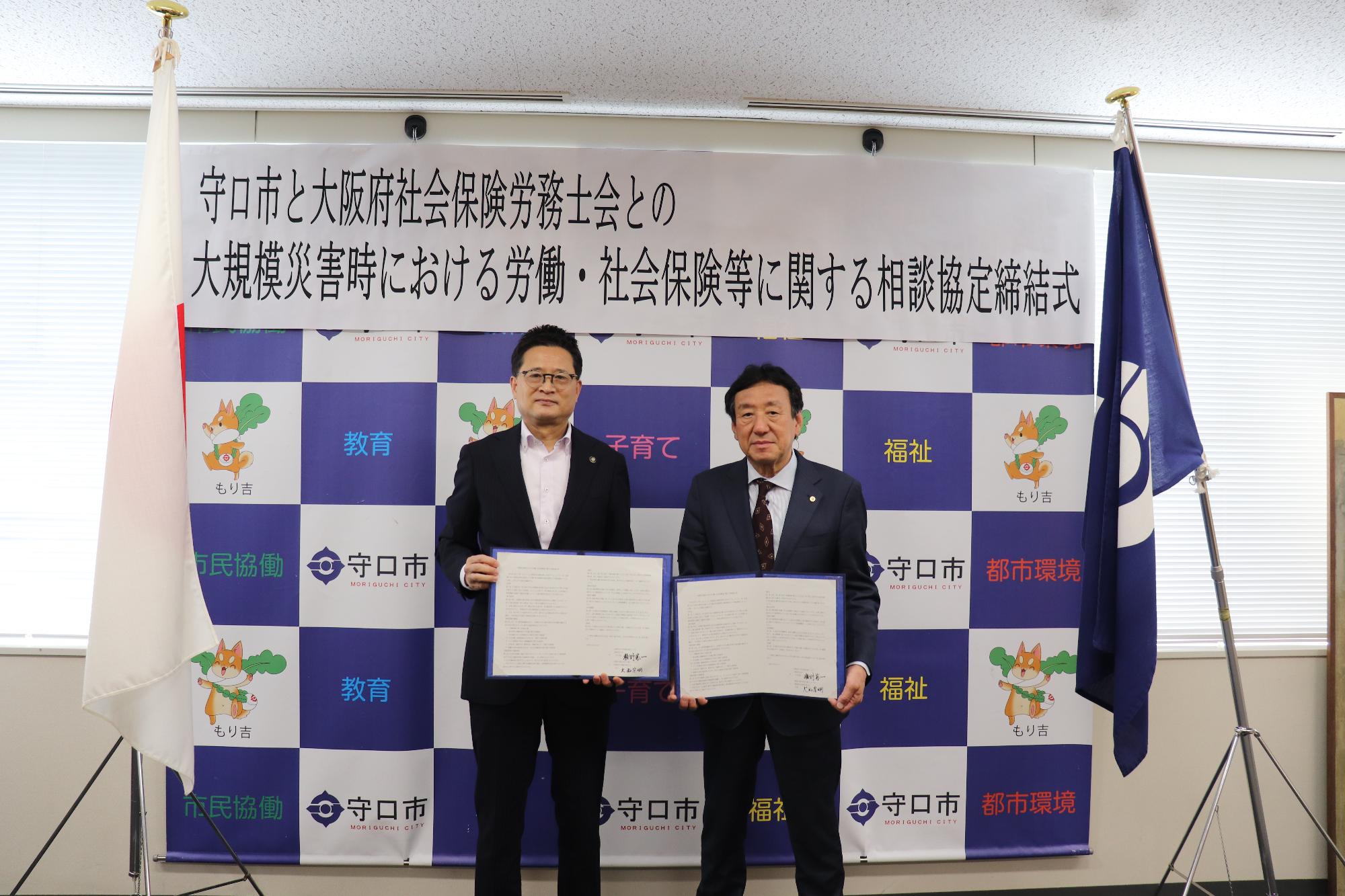 椅子に座り、正面を向き二つ折りの書類を開くスーツ姿の男性と、その左隣に設置されたモニターに映る一人の男性の写真