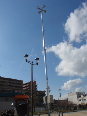 広い敷地に街頭と並んで設置された高いポールの先にスピーカーが4つ付いた防災無線の全体の写真