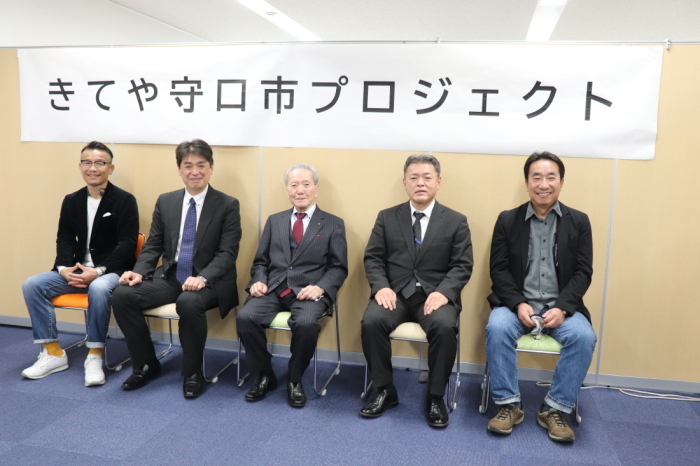 「きてや守口市プロジェクト」と書かれた横断幕の下に関係者5名の男性が横一列に並んで椅子に座っている写真