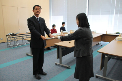 スーツ姿の男性が、グレーのスーツを着た女性に選考結果を手渡ししている写真