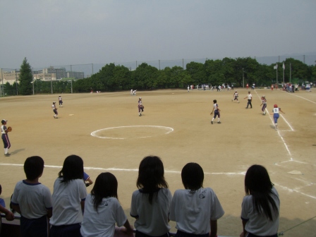 グラウンドでソフトボールの試合が行われている様子を見ている6名の女子生徒たちの後ろ姿の写真