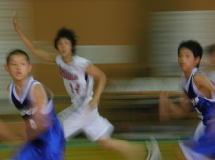 青いユニフォームを着た選手2名と、右手を上にあげている白いユニフォームを着た選手1人がコートの中を走っているバスケットボールの試合の写真