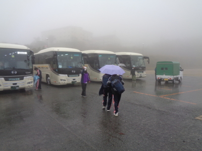 雨の中を女子生徒二人が一つの傘に入りながら、奥に停まっているバスの方へ歩いている写真