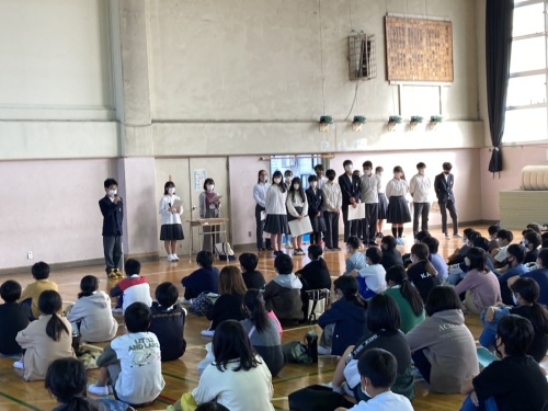 体育館で体育座りしている小学生が見ているなか複数の中学生が前に立って話をしているオープンスクールの様子の写真