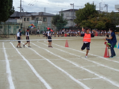 オレンジのビブスを着た男子生徒がリレーのバトンを手にゴールテープを走り抜けようとする瞬間の写真