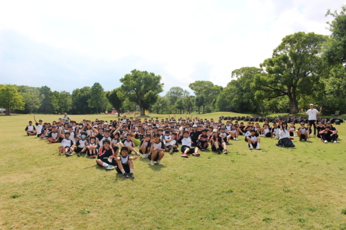 木々に囲まれた芝生広場に校外学習に訪れた生徒たちが体育座りしてカメラに向かってピースサインをしている写真