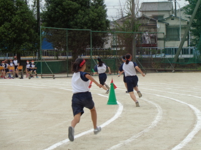 校庭のトラックの中心側を走るバトンを持った4人の女子生徒を後方から写した写真