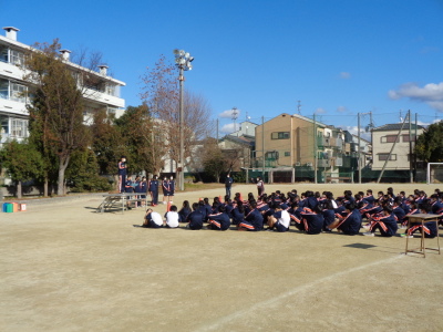 校庭に体育座りしている生徒たちが檀上に立って話をしている生徒の話を聞いている写真