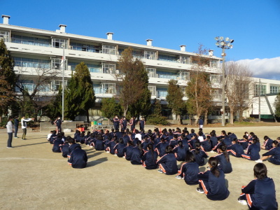 運動場に座っている生徒たちの前に代表の4名の生徒が立っている写真