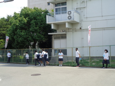 校舎横にあるフェンスの前に立って生徒たちが広がらない運動で通学中の生徒に声掛けしている様子の写真