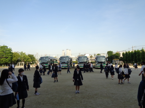 校庭のグラウンドに4台のバスが停車しており、制服を着た学生が集まったり、バスの方へ歩いている写真