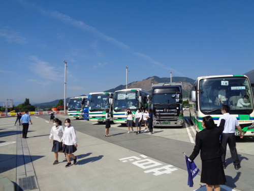 青空の下、停車するバスの周りにいる学生や旗を持つ添乗員さんの後ろ姿の写真