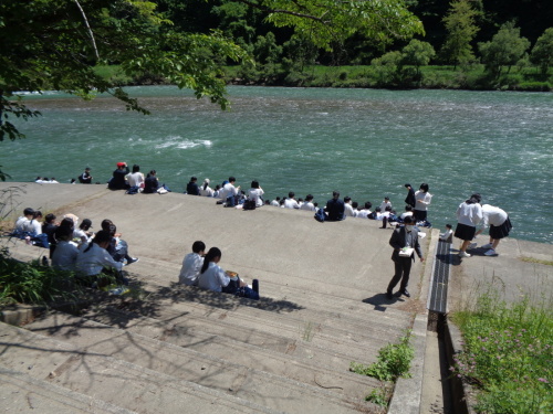 流れる川の前の階段に座っている生徒たちの後ろ姿の写真