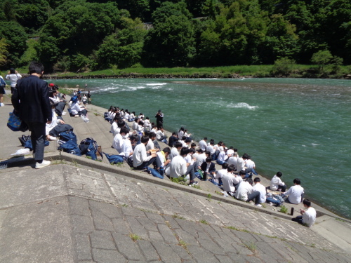 流れが強い川の前の階段で、学生たちが座って手元を見ている写真