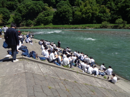 流れが強い川の前の階段で、学生たちが座って手元を見ている写真
