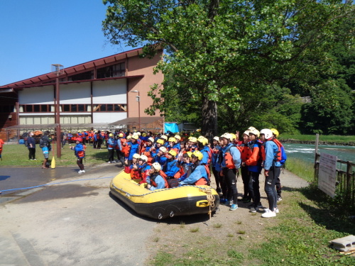 黄色いゴムボートの上で、オレンジの救命胴衣をつけ、ウェットスーツに青いジャケットを着た学生が記念写真をしている写真