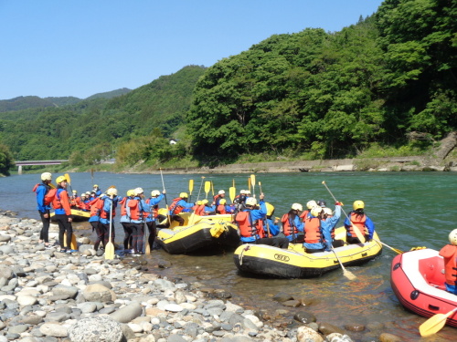 救命胴衣やウェットスーツを着た学生が、黄色いゴムボートの上とその周りに、パドルを持って集まっている写真