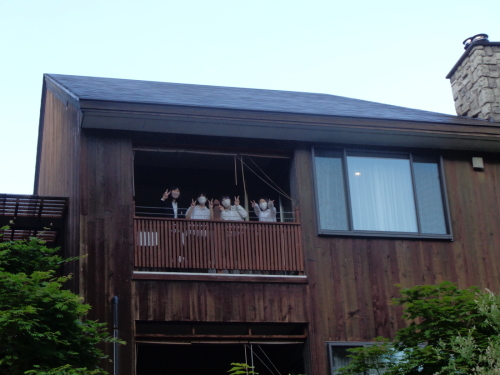 三角屋根で木目調外壁の建物の2階バルコニーで、4名の学生たちがピースサインをしている写真