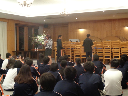 座って並んでいる学生が注目する中、ストライプシャツに黒いスカートを着た学生と、2名の学生が後ろをむいて立っている写真