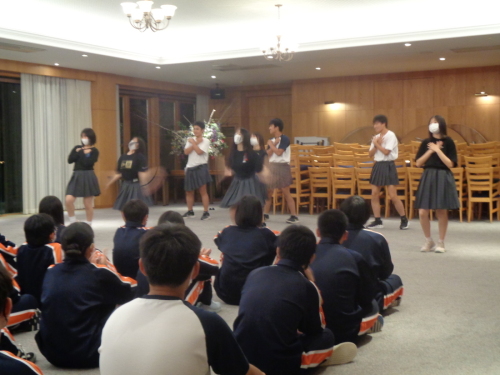 グレーのスカートに黒いシャツを着た女子学生と、グレーのスカートに白いシャツを着た男子学生が踊っている様子に注目している学生達の写真