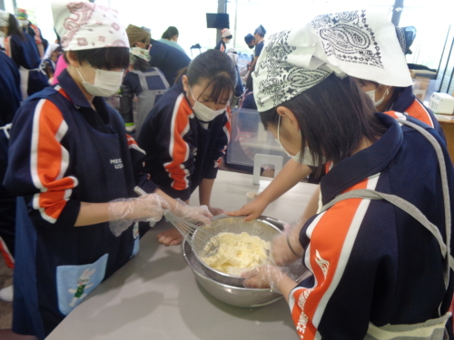 三角巾にエプロンを付けた学生が、泡だて器を使いボールの中の薄黄色の食材を混ぜている写真