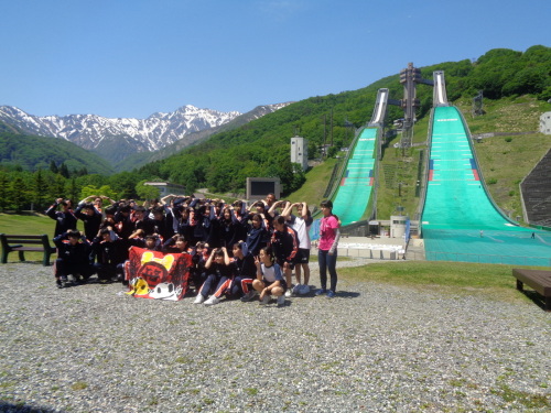 赤い生地に絵が描かれた旗を持って、緑のジャンプ台の前でポーズをとって記念撮影をしている学生たちの写真