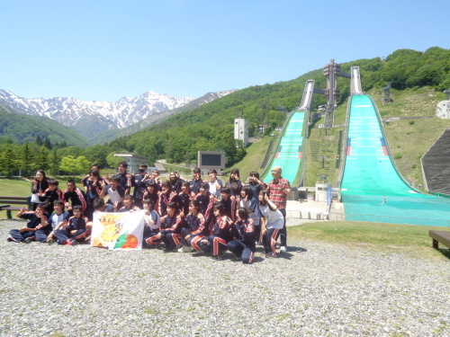 白い生地に絵が描かれた旗を持って、緑のジャンプ台の前でポーズをとって記念撮影をしている学生たちの写真
