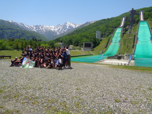 青と緑のような色の旗を持って、緑のジャンプ台の前でポーズをとって記念撮影をしている学生たちの写真