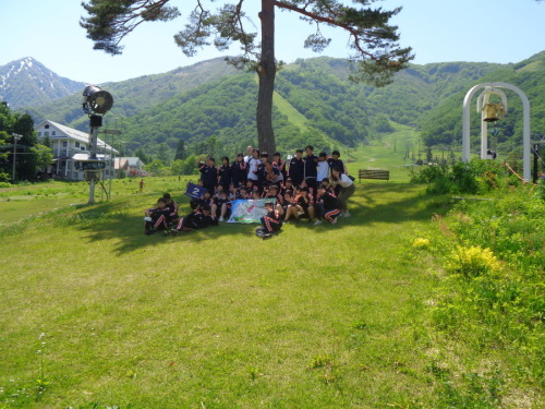 大きな木の下の芝生で、薄い青色の旗をもった学生達が集まり記念撮影をしている写真