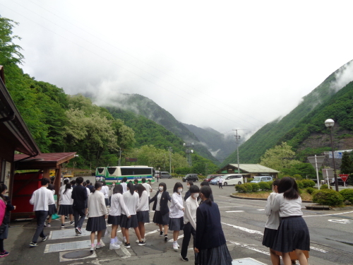 学生が歩き出す先の駐車場にバスが駐車していて、奥の山に雲がかかっている写真