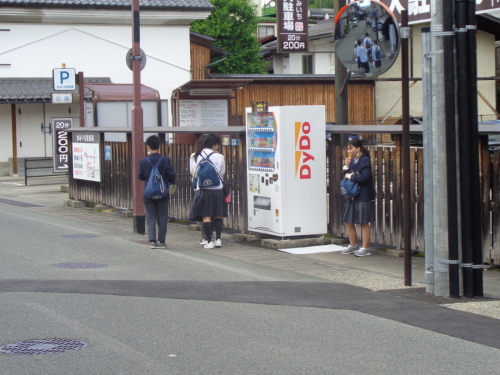 駐車場の横に設置された自動販売機の近くに立つ4名の男女学生の写真