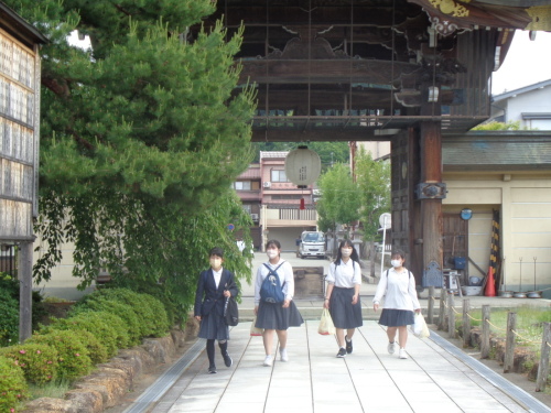 大きな門を後ろに、4名の女子学生が横に並んで歩いている写真