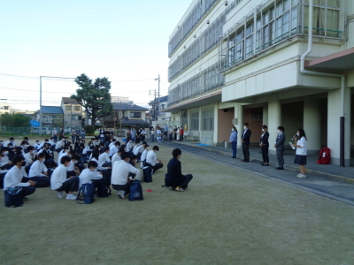 校舎前に生徒達が整列して座り、先生方の話を聞いている様子の写真