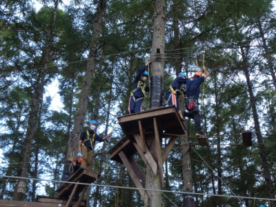 高い木の上に足場が作られ、木と木の間に板とロープで作られた橋があり、安全ベルトのワイヤーを持ちながら移動しようとしている生徒達の写真