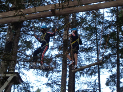 ヘルメットと安全ベルトを付けた生徒2名が高い木の上に造られた丸太の橋を渡っている写真