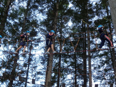 高い木と木の間にロープで作られた橋を3名の生徒が渡っている写真
