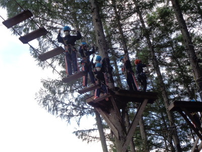 高い木と木の間に間隔を空けて置かれた板とローブで作られた橋を、生徒達が順番に渡っている様子を左下から見上げるように写した写真