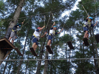 高い木と木の間に、間隔を空けて置かれた板とロープで作られた橋の板の上に、生徒達が安全ベルトのワイヤーを握りながら移動している写真