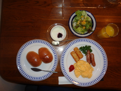 お皿に盛り付けられたロールパン、スクランブルエッグとウインナー、ポテト、サラダ、ヨーグルト、オレンジジュースの写真