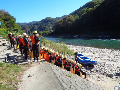 ヘルメットと救命胴衣を身に着けた生徒達が、河原の堤防の階段に整列して座っている写真