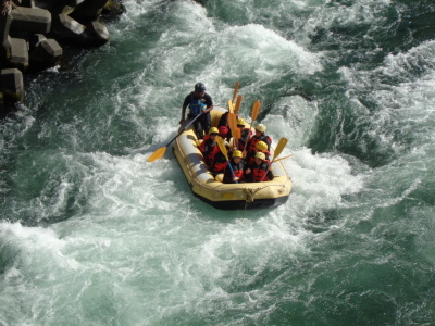 濁流の中で生徒達がオールを上に持ち上げ、船頭者の男性が、オールでボートの向きを変えて川を下っている様子の写真