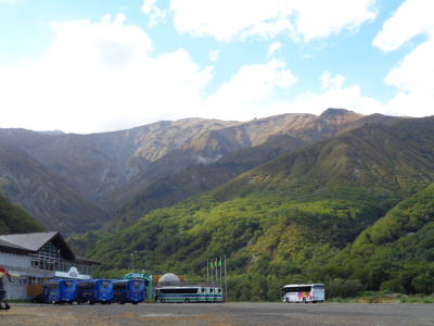 山のふもとにある旅行先の敷地内から、バスが並んでいる駐車場や奥に連なる山並みを写した写真