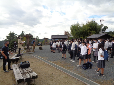 砂利の敷かれた敷地で前に立って話をする先生と、話を聞いている生徒たちの写真