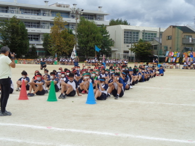 校庭の中心に赤と緑と青のカラーコーンが置いてあり、置いてあるカラーコーンの色に合わせて生徒たちが縦1列に並んで座っている写真