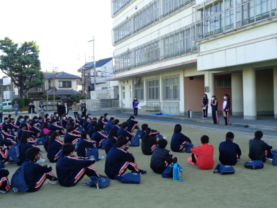 ジャージ姿の生徒たちが校舎の前の運動場に座っている写真