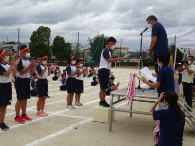 校庭の演台に立っている先生が、オレンジ色の鉢巻をした男子生徒に表彰状を渡している写真